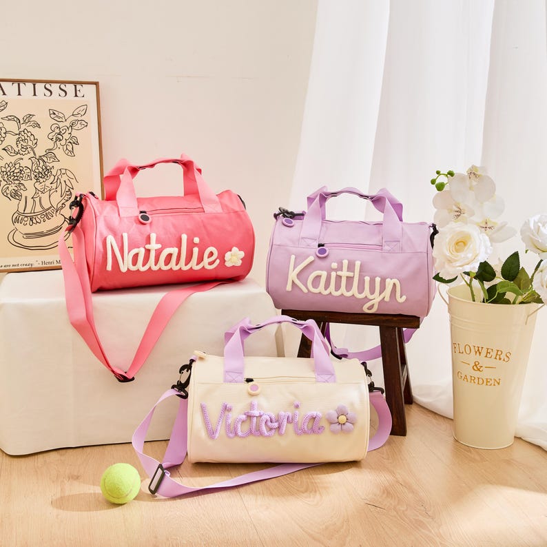 May include: Three personalized duffel bags in pink, lavender, and cream. Each bag features a name embroidered in white script with a floral accent. The bags have top handles and adjustable shoulder straps, perfect for carrying sports gear or travel essentials.