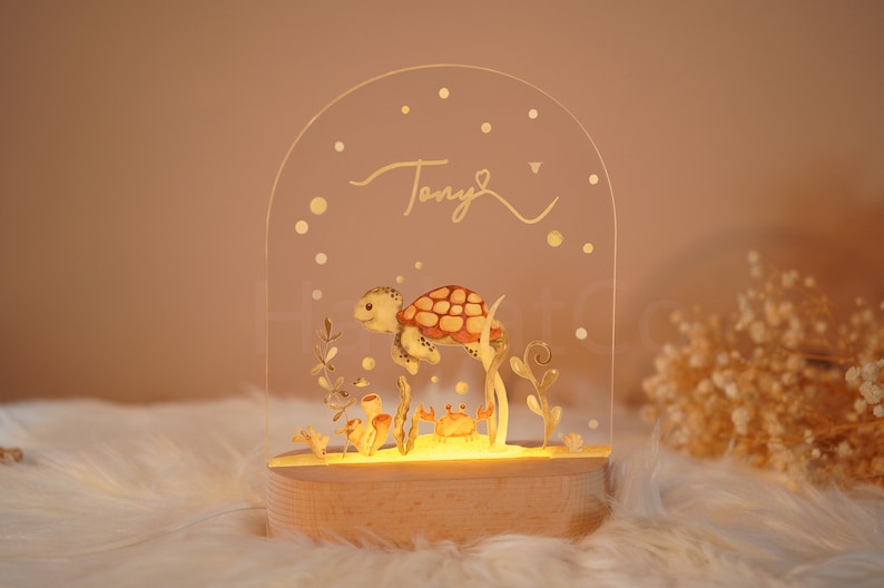 Op de afbeelding: Een helder acryl nachtlampje met een houten voet. Het lampje heeft een handgeschilderde illustratie van een schildpad die in de oceaan zwemt met zeewier en koraal. De naam "Tony" staat in gouden letters bovenaan het lampje.