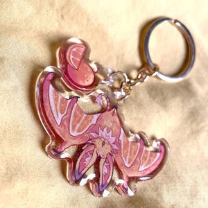 May include: A clear acrylic keychain featuring a stylized bat design with pink, orange, and purple hues. The bat has large ears and outstretched wings, with glitter accents. The keychain includes a silver-colored keyring and chain.