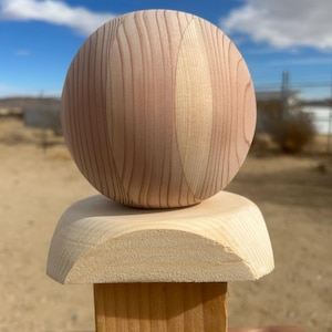 May include: A wooden sculpture featuring a light brown sphere atop a light-colored, curved base, which is mounted on a square post. The wood grain is visible, and the sculpture is set against a backdrop of a blue sky.