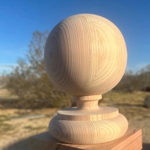 May include: A wooden finial with a spherical top and a decorative base. The finial is made of light-colored wood and is mounted on a square wooden post. The background features a clear blue sky and natural outdoor setting.