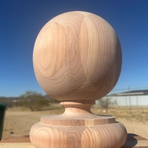 May include: A light-colored, unfinished wooden finial with a round top and a decorative base. The finial is made of multiple pieces of wood, with visible grain patterns. The background features a clear blue sky and a natural outdoor setting.