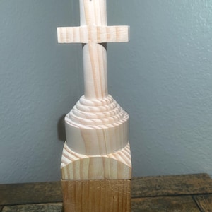 May include: A wooden cross sculpture with a light brown finish. The cross is mounted on a carved, layered base. The sculpture is made of wood and has a natural, unfinished appearance.