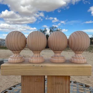 May include: Four unfinished wooden finials with a ball-shaped top and a decorative base, arranged on a wooden plank. The finials are made of light-colored wood and have a textured, grooved design. The background features a blue sky with fluffy white clouds.