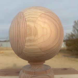 May include: A wooden decorative sphere with a carved design, mounted on a matching wooden base. The sphere is light brown with visible wood grain and curved lines. The base has a classic, rounded shape. The piece is displayed outdoors.