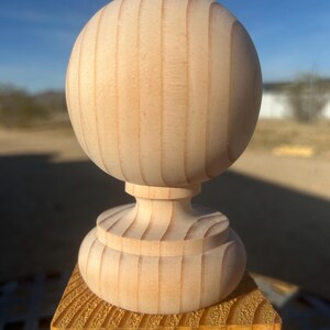 May include: A wooden finial with a spherical top and a decorative base. The finial is made of unfinished wood and is mounted on a square wooden post. The wood grain is visible, and the piece is outdoors in natural light.