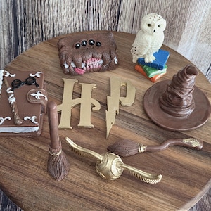 May include: A collection of miniature fantasy-themed items including a brown book with glasses, a furry brown monster book, a white owl on colourful books, a brown sorting hat, a gold HP logo, a broomstick, and a gold winged ball figurine.