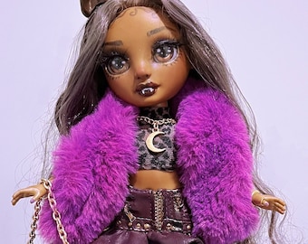 Può includere: Una bambola di moda con lunghi capelli castani, che indossa un cappotto di pelliccia viola, un top nero e oro, una gonna nera e stivali neri con dettagli dorati. La bambola tiene una borsa viola con una catena dorata.