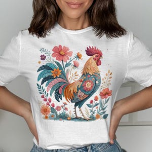 以下が含まれることがあります： 花柄のカラフルな雄鶏のデザインが施された白いTシャツ。雄鶏は赤い鶏冠、オレンジと黄色の羽、体に青い花柄があります。花はピンク、オレンジ、青です。