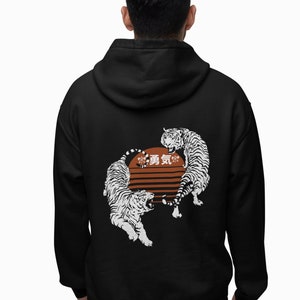 May include: Black hoodie featuring a graphic of two white tigers facing each other in front of a brown and orange sun design. Japanese characters are above the sun. The hoodie has a hood and long sleeves.