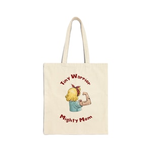 May include: Cream-colored tote bag with a cartoon illustration of a woman flexing her arm, wearing a red bandana. The text "Tiny Warrior" and "Mighty Mom" are printed in maroon. The bag has two handles.