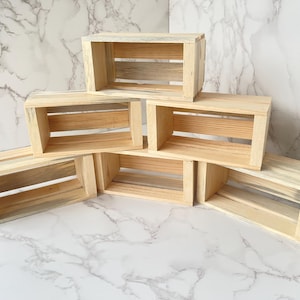 May include: Six small, unfinished wooden crates are stacked in a visually appealing arrangement. The crates are rectangular with open fronts and backs, and the wood grain is visible. The crates are made of light-colored wood.