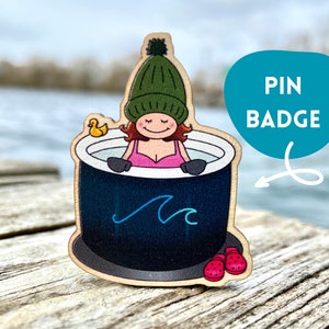 May include: A wooden pin badge featuring a cartoon illustration of a person wearing a green knitted hat and pink swimsuit relaxing in a hot tub with a rubber ducky. The hot tub is black with a blue wave design and the text "PIN BADGE" is written in a blue circle.