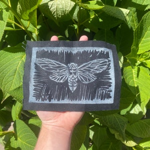 May include: A black patch with a white cicada insect design. The insect is centered on the patch, with detailed wings and body. The patch is rectangular with a white border and is held against a background of green leaves.
