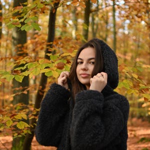 Puede incluir: Una mujer con una chaqueta negra con capucha y textura peluda se encuentra en un bosque con follaje oto&ntilde;al. Las hojas son principalmente amarillas y naranjas.