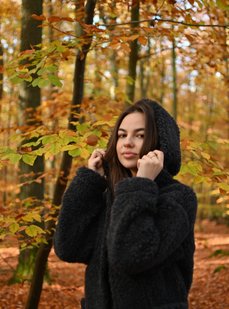 Puede incluir: Una mujer con una chaqueta negra con capucha y textura peluda se encuentra en un bosque con follaje oto&ntilde;al. Las hojas son principalmente amarillas y naranjas.