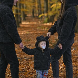 Puede incluir: Una familia de tres personas con chaquetas con capucha de pelo negro a juego camina por un bosque con hojas ca&iacute;das. El ni&ntilde;o lleva un chupete y tiene orejas de oso en la capucha.