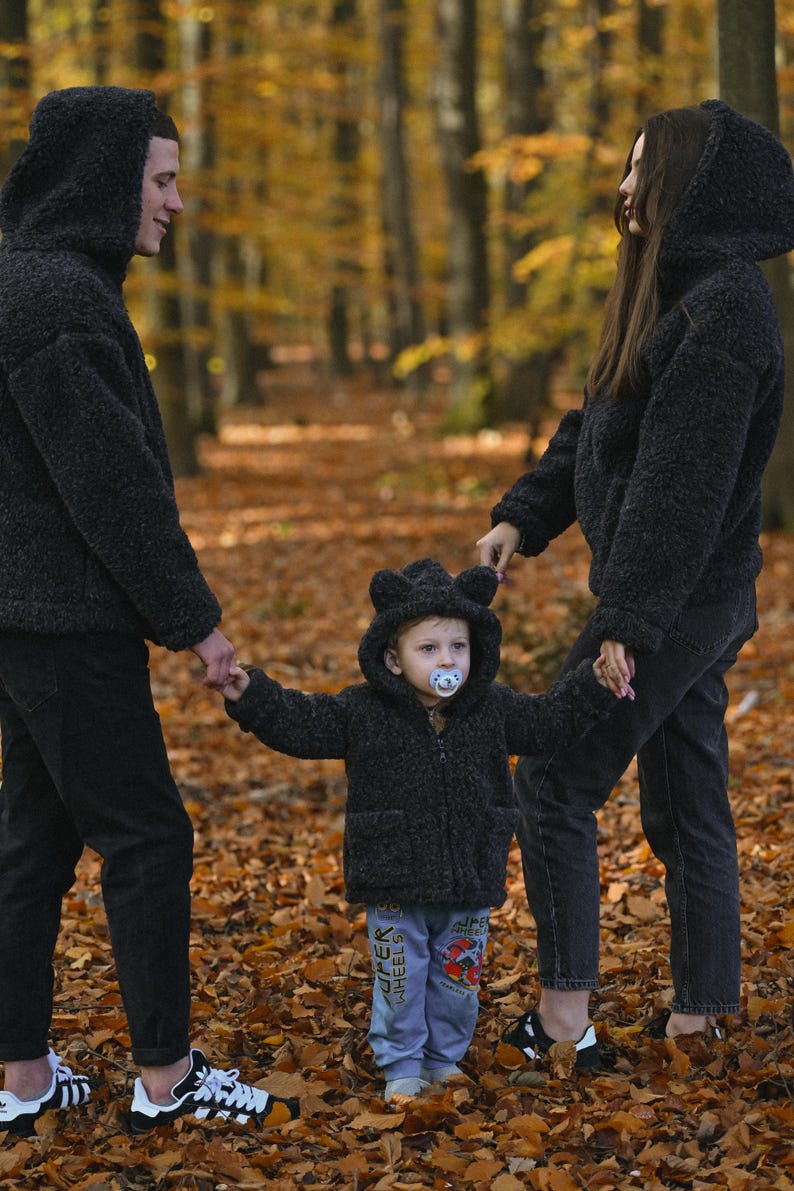 Puede incluir: Una familia de tres personas con chaquetas con capucha de pelo negro a juego camina por un bosque con hojas ca&iacute;das. El ni&ntilde;o lleva un chupete y tiene orejas de oso en la capucha.