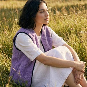 K&ouml;nnte beinhalten: Eine Person tr&auml;gt eine lavendelfarbene Fleeceweste mit schwarzem Besatz &uuml;ber einem wei&szlig;en Kleid und sitzt in einem Feld mit hohem Gras. Die Weste ist aus weichem, strukturiertem Material. Die Szene ist von warmem Sonnenlicht durchflutet.