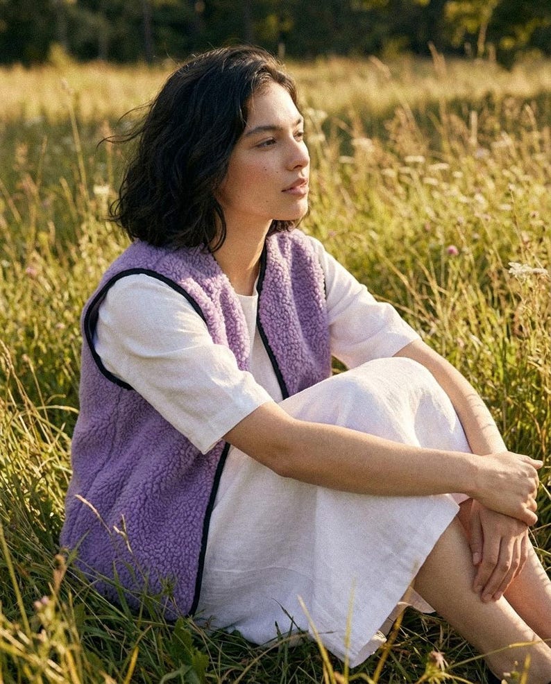 K&ouml;nnte beinhalten: Eine Person tr&auml;gt eine lavendelfarbene Fleeceweste mit schwarzem Besatz &uuml;ber einem wei&szlig;en Kleid und sitzt in einem Feld mit hohem Gras. Die Weste ist aus weichem, strukturiertem Material. Die Szene ist von warmem Sonnenlicht durchflutet.