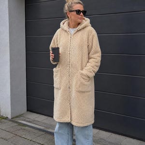 Op de afbeelding: Een lange, beige teddy fleece jas met capuchon, ritssluiting aan de voorzijde en twee grote opgestikte zakken. De jas wordt gedragen over een lichtblauwe wijde spijkerbroek en witte sportschoenen. Een zwarte beker wordt in de hand gehouden.
