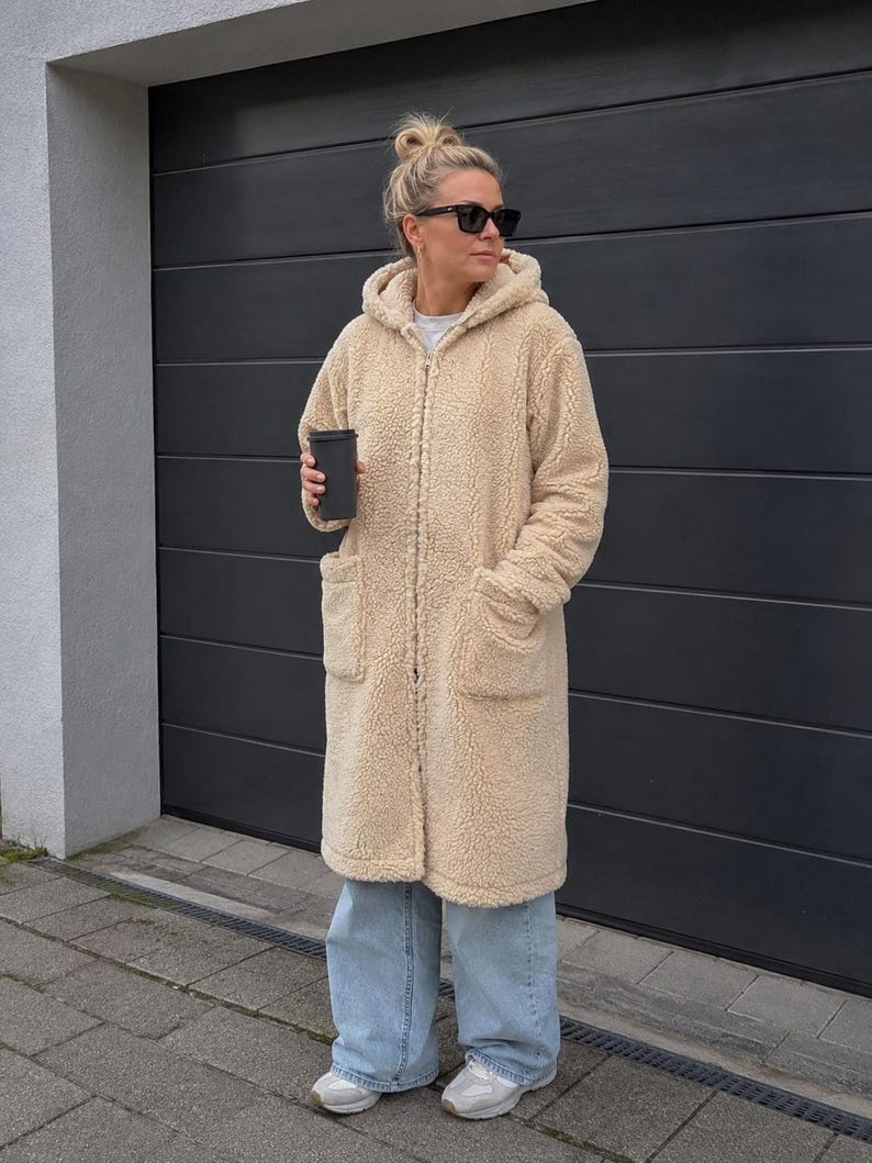 May include: A long, beige, hooded teddy fleece coat with a front zipper closure and two large patch pockets. The coat is worn over light blue wide-leg denim jeans and white athletic sneakers. A black cup is held in one hand.