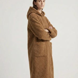 May include: A long, brown, textured teddy fleece coat featuring a hood, long sleeves, and two large front patch pockets. The coat is constructed from a thick, fuzzy material with a visible pile, extending to mid-thigh length with a straight hem.