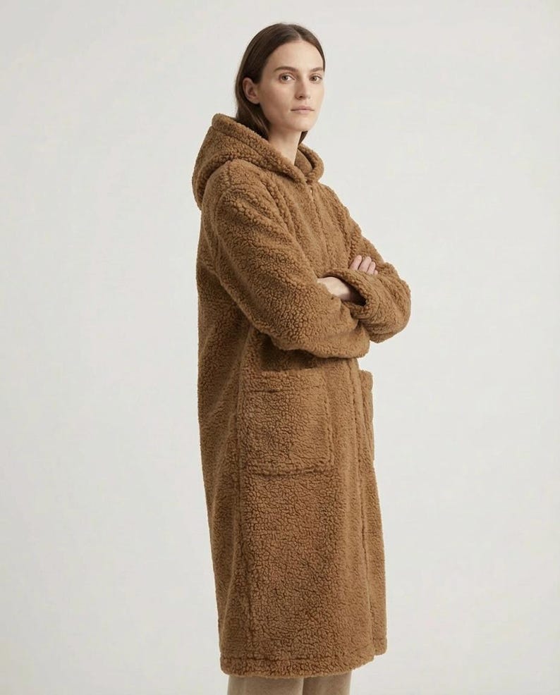 May include: A long, brown, textured teddy fleece coat featuring a hood, long sleeves, and two large front patch pockets. The coat is constructed from a thick, fuzzy material with a visible pile, extending to mid-thigh length with a straight hem.