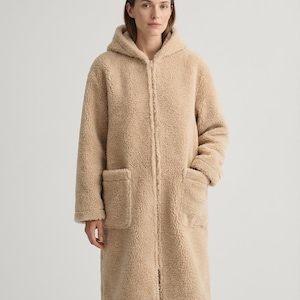 May include: A long, beige, hooded coat made of a textured, fleece-like material. The coat features a full-length front zipper closure, long sleeves, and two large, rectangular patch pockets on the front, extending to the mid-thigh area.