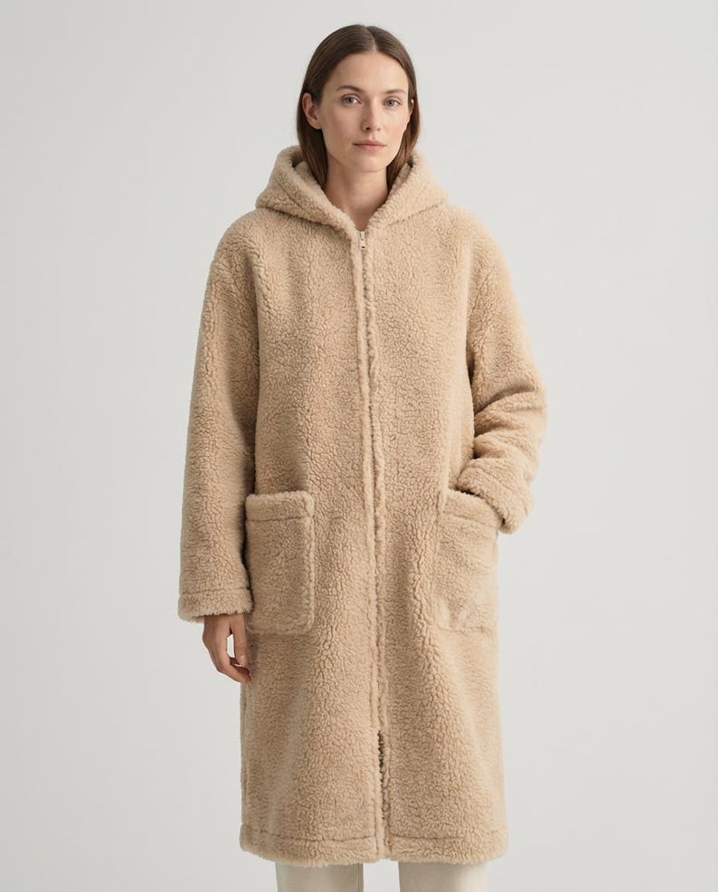 May include: A long, beige, hooded coat made of a textured, fleece-like material. The coat features a full-length front zipper closure, long sleeves, and two large, rectangular patch pockets on the front, extending to the mid-thigh area.