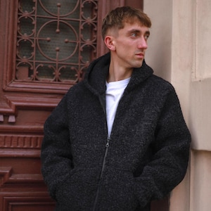Könnte beinhalten: Ein dunkelgrauer, texturierter Kapuzenpullover mit Reißverschluss, Kapuze und zwei Vordertaschen, getragen über einem schlichten weißen T-Shirt. Das Kleidungsstück besteht aus einem dicken, flauschigen, fleeceartigen Material mit sichtbarer Strickstruktur.