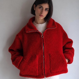 Puede incluir: Una chaqueta de forro polar de color rojo brillante, de manga larga y textura esponjosa, con cuello alto, cremallera metálica plateada de longitud completa con tirador circular y dos grandes bolsillos de parche en la parte delantera.