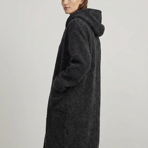 May include: A long, charcoal gray coat made of a textured, fuzzy fleece material. The coat features a hood, long sleeves, and two side pockets. The garment has a straight, knee-length silhouette and is worn over dark pants.