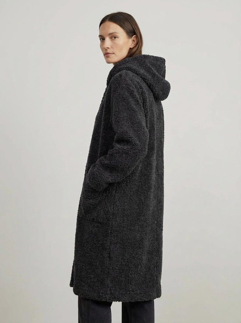 May include: A long, charcoal gray coat made of a textured, fuzzy fleece material. The coat features a hood, long sleeves, and two side pockets. The garment has a straight, knee-length silhouette and is worn over dark pants.