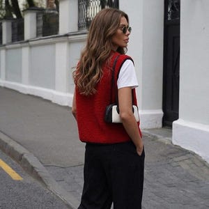 Puede incluir: Una persona con un chaleco rojo sobre una camiseta blanca y pantalones negros. La persona tambi&eacute;n lleva gafas de sol y lleva un peque&ntilde;o bolso blanco y negro. El fondo incluye una pared blanca y una puerta negra.