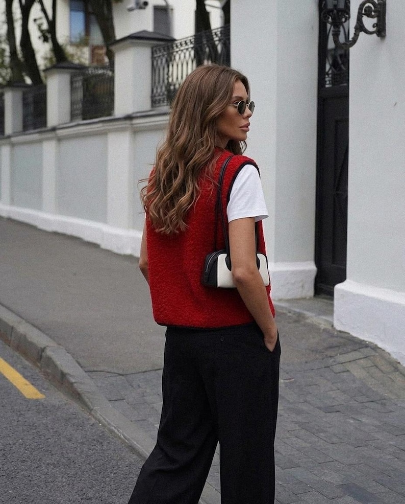 Puede incluir: Una persona con un chaleco rojo sobre una camiseta blanca y pantalones negros. La persona tambi&eacute;n lleva gafas de sol y lleva un peque&ntilde;o bolso blanco y negro. El fondo incluye una pared blanca y una puerta negra.
