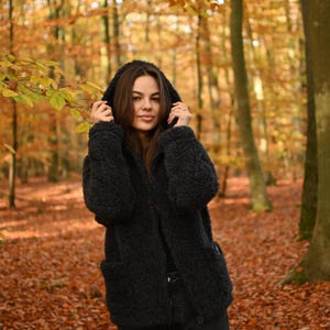 Puede incluir: Una mujer con una chaqueta de forro polar negra con capucha en un entorno de bosque con follaje oto&ntilde;al.