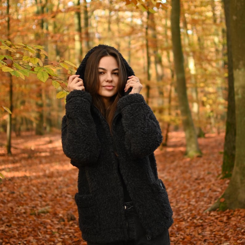 Puede incluir: Una mujer con una chaqueta de forro polar negra con capucha en un entorno de bosque con follaje oto&ntilde;al.