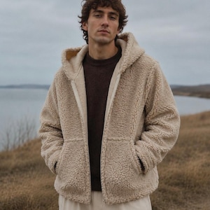Könnte beinhalten: Ein beigefarbener Kapuzenpullover mit Reißverschluss aus Sherpa-Fleece mit strukturierter, lockiger Oberfläche. Das Kleidungsstück verfügt über einen durchgehenden Frontreißverschluss, eine Kapuze und zwei Vordertaschen. Er wird über einem dunkelbraunen Pullover getragen.