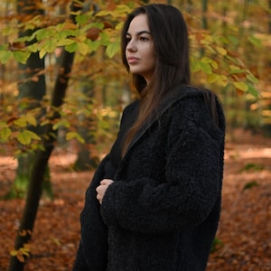 Puede incluir: Una mujer con una chaqueta peluda negra se encuentra en un bosque con follaje oto&ntilde;al. Est&aacute; mirando hacia un lado.