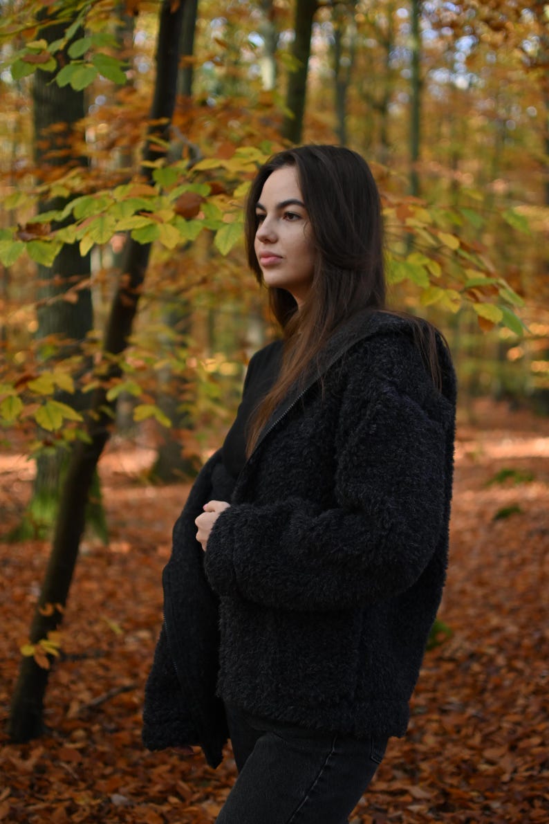 Puede incluir: Una mujer con una chaqueta peluda negra se encuentra en un bosque con follaje oto&ntilde;al. Est&aacute; mirando hacia un lado.