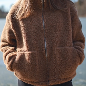Puede incluir: Chaqueta de forro polar marrón con cuello alto y cremallera completa. La chaqueta tiene dos bolsillos delanteros y un tirador de cremallera en forma de anillo. El modelo lleva pantalones negros. La chaqueta es de un material suave y texturizado.
