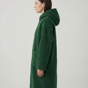 May include: A long, forest green coat made of a textured, curly fleece material. The coat features a hood, long sleeves, and a straight-cut silhouette that reaches below the hips. The design includes a visible side pocket on the front panel.