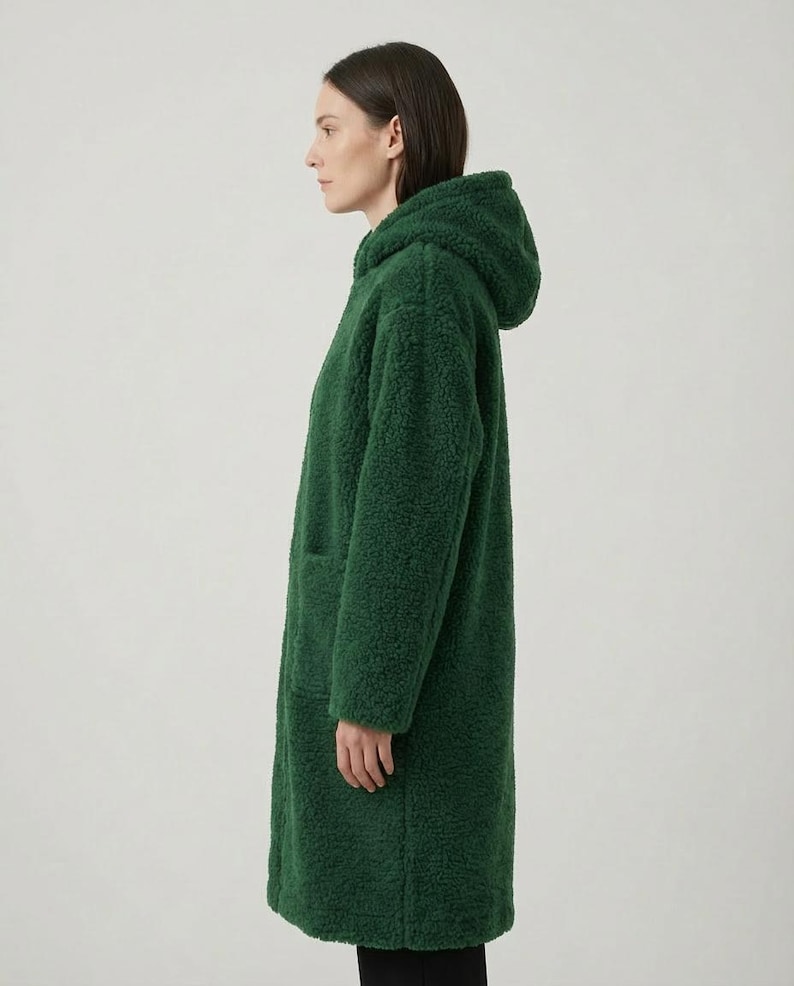 May include: A long, forest green coat made of a textured, curly fleece material. The coat features a hood, long sleeves, and a straight-cut silhouette that reaches below the hips. The design includes a visible side pocket on the front panel.