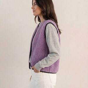 K&ouml;nnte beinhalten: Eine Frau tr&auml;gt eine lavendelfarbene Fleeceweste mit schwarzem Besatz &uuml;ber einem grauen Langarmshirt und einer wei&szlig;en Hose. Die Weste hat ein weiches, strukturiertes Aussehen. Das Outfit ist l&auml;ssig und bequem.