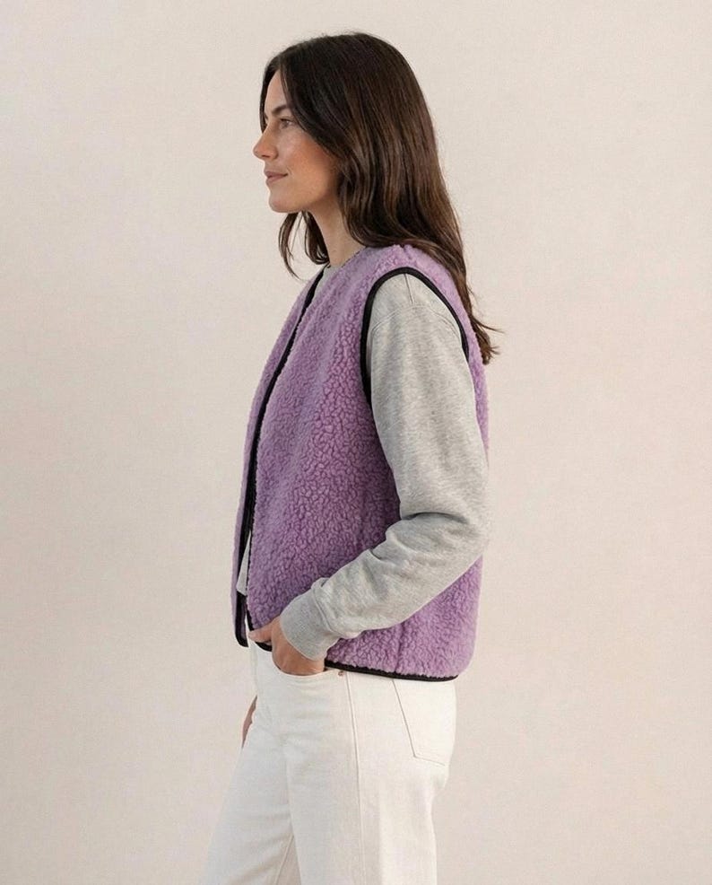 K&ouml;nnte beinhalten: Eine Frau tr&auml;gt eine lavendelfarbene Fleeceweste mit schwarzem Besatz &uuml;ber einem grauen Langarmshirt und einer wei&szlig;en Hose. Die Weste hat ein weiches, strukturiertes Aussehen. Das Outfit ist l&auml;ssig und bequem.