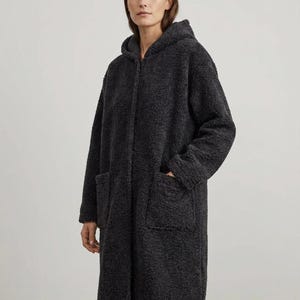May include: A charcoal gray long-length coat made of a textured, fuzzy fleece material. The coat features a hood, long sleeves, and two large patch pockets on the front. It has a straight silhouette and a zip-up front closure that is concealed.