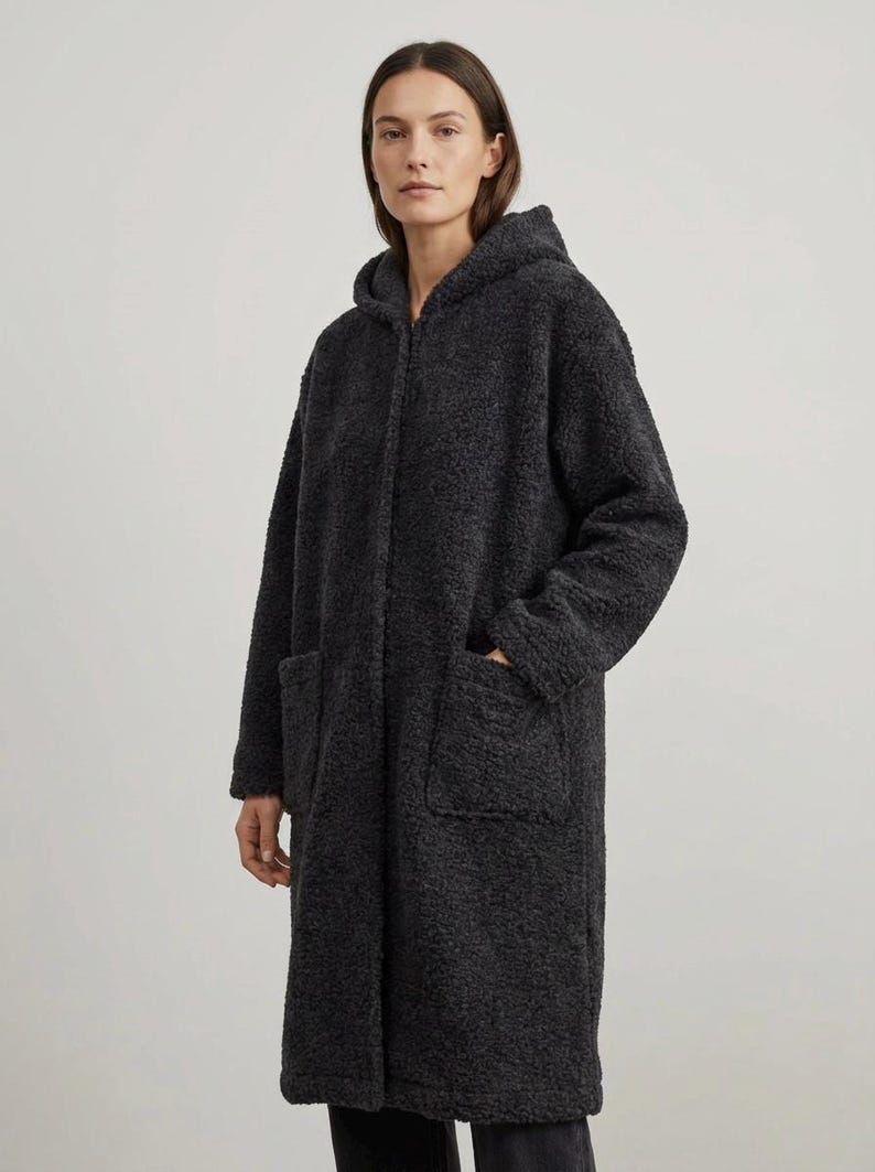 May include: A charcoal gray long-length coat made of a textured, fuzzy fleece material. The coat features a hood, long sleeves, and two large patch pockets on the front. It has a straight silhouette and a zip-up front closure that is concealed.