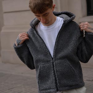 Peut inclure: Un sweat à capuche zippé gris anthracite en matière polaire texturée et duveteuse. Le vêtement présente une capuche, une fermeture éclair intégrale sur le devant et deux grandes poches avant. Il est porté sur un t-shirt blanc à col rond.