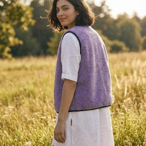 K&ouml;nnte beinhalten: Eine Frau tr&auml;gt eine lavendelfarbene Fleeceweste mit schwarzem Besatz &uuml;ber einem wei&szlig;en Kurzarmshirt und einem wei&szlig;en Rock. Sie steht in einem Feld mit hohem Gras, im Hintergrund B&auml;ume. Die Weste ist das zentrale Kleidungsst&uuml;ck.